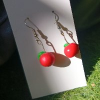 Handcrafted polymer clay dangle earring shaped like a glossy red apple with a tiny green leaf on a silver hook, worn by a woman and displayed on a white card.
