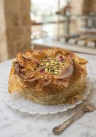 עוגת צ'יזקייק בקלאווה ופיסטוק גבוהה ושלמה של הפטיסרי של חיים בירושלים, עטופה בכתר בצק פילו פריך וזהוב ומקושטת בהמון פיסטוקים קלויים.