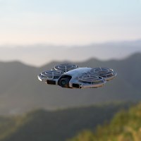 רחפן DJI Neo 2 Fly More Combo – פריט בודד אפור בהיר עם מגיני מדחפים טס באוויר מעל נוף הררי