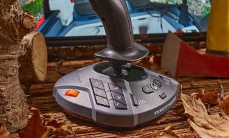 בקר טיסה Thrustmaster SimTask Farmstick Xbox/PC – הבקר האפור-שחור מונח על משטח עץ עם עלים יבשים כשמסך משחק וגרזן נראים ברקע
