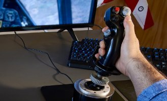 בקר טיסה Thrustmaster F-16C Viper Strike Fighter PC – יד אוחזת בבקר טיסה שחור על שולחן מול מסך מחשב פעיל