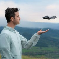 רחפן ללא שלט DJI Neo 2 Fly More Combo – גבר מביט בכלי טיס כסוף מרחף מעל כף ידו על רקע נוף ירוק