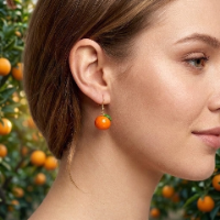 Handcrafted polymer clay dangle earring shaped like a glossy orange with a tiny green leaf on a gold hook, worn by a woman with an orange grove in the background.